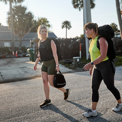 Two women stroll down a sunny street, each with gym bags in tow, their outfits complemented by functional fitness footwear. Palm trees and a fence frame the backdrop as they engage in lively conversation, their GORUCK Women's Ballistic Trainers in Ranger Green + White W / Coyote Reflective Spearhead providing comfort with every step thanks to the 3X Support™ technology.