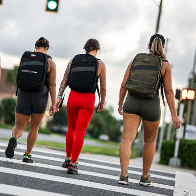 Three people sporting backpacks stride across a crosswalk. The individual on the left carries a CORDURA® Ballistic Nylon backpack, the one in the center wears red leggings, and the one on the right boasts GORUCK Women's Ballistic Trainers - Ranger Green + White with Coyote Reflective Spearhead. Traffic lights glow in the background.