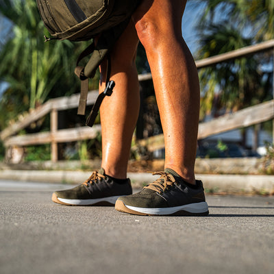 A close-up shows someone wearing the Women's Ballistic Trainers by GORUCK in Ranger Green and White with Coyote Reflective Spearhead, designed as functional fitness footwear with 3X Support™. They stand on a sunlit pavement carrying a green backpack, while blurred palm trees and a wooden fence are visible in the background.