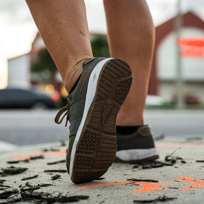 A close-up shot captures a person walking on a sidewalk wearing GORUCK's Women's Ballistic Trainers in Ranger Green and White, accented with Coyote Reflective Spearhead. Constructed with CORDURA® Ballistic Nylon, these sneakers feature white soles. The foot is caught mid-step among scattered leaves, with blurred buildings and cars in the background.