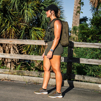 Clad in athletic gear, a person wearing a green top and shorts carries a CORDURA® Ballistic Nylon backpack as they stand on a path surrounded by lush greenery and palm trees. It's a bright, sunny day, and they sport GORUCK's Women's Ballistic Trainers in Ranger Green with White and Coyote Reflective Spearhead for functional fitness mobility, complemented by a cap for extra sun protection.