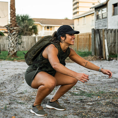 A person wearing a cap, tank top, shorts, and the GORUCK Women's Ballistic Trainers in Ranger Green + White with Coyote Reflective Spearhead is squatting outdoors with a backpack on. They are in a sandy area with a wooden fence and houses in the background, giving the setting a casual and relaxed vibe.