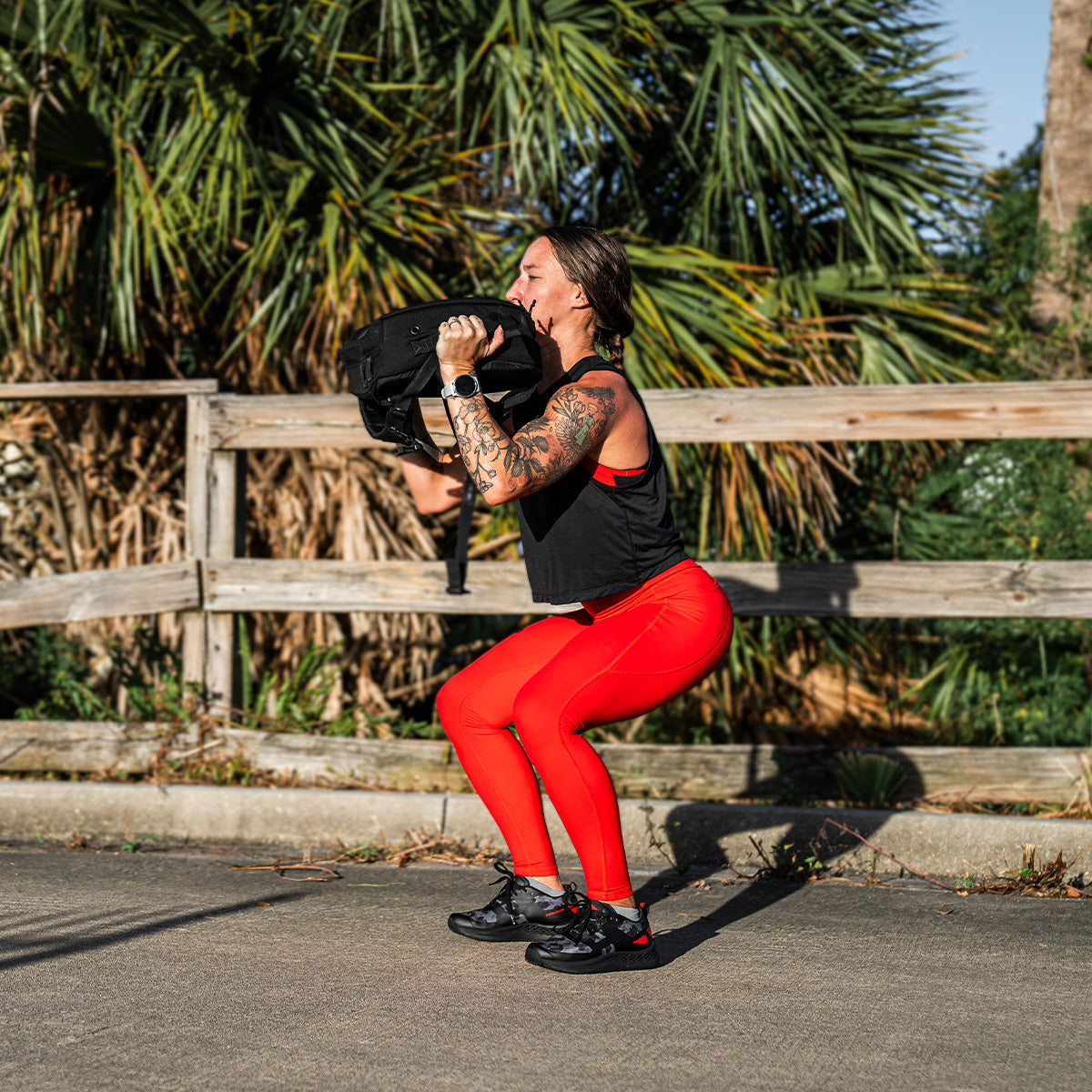 A person is exercising outdoors, performing a squat while holding a black sandbag. They are wearing GORUCK's Women's Rough Runner in Midnight Frogskin and High Risk Red, which features a black tank top paired with vibrant red leggings ideal for any Rough Runner. A wooden fence and green foliage provide the perfect backdrop for this dynamic scene.