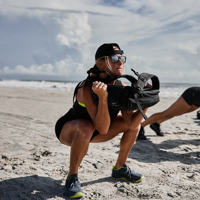 A woman in sunglasses and a cap performs a sandbag squat on the beach, highlighting her Women's Ballistic Trainers - Wolf Grey + Acid Lime W / Acid Lime Reflective Spearhead by GORUCK. Her black tank top and shorts complement her neon-accented sneakers. Another person is partially visible against the backdrop of a cloudy sky.