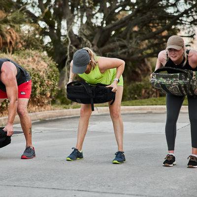 Three individuals are exercising outdoors, bending forward while holding weighted bags made from CORDURA® Ballistic Nylon. They are dressed in athletic clothing, including caps, shorts, leggings, and wearing GORUCK's Women's Ballistic Trainers in Wolf Grey and Acid Lime with Acid Lime Reflective Spearhead. Trees and bushes can be seen in the background.