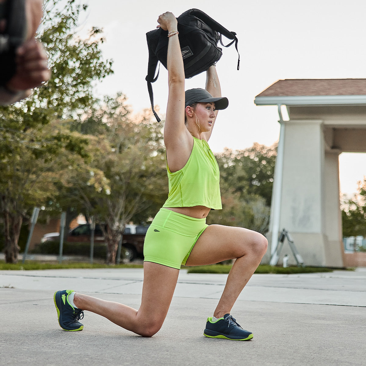 Clad in Wolf Grey and Acid Lime Women's Ballistic Trainers by GORUCK, a person performs lunges on a concrete surface, effortlessly lifting a backpack overhead. Their functional fitness footwear ensures smooth movement, while trees and a white structure provide a serene backdrop.