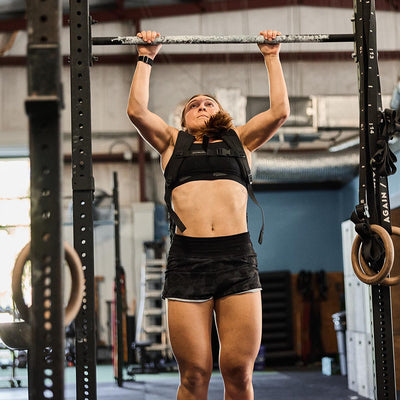 A woman performs a pull-up in a gym, sporting GORUCK’s ToughStretch Women's Training Shorts and a black athletic top. She utilizes a pull-up bar with gymnastic rings visible on either side. The gym is equipped with industrial-style equipment and lighting, showcasing her dedication to fitness.