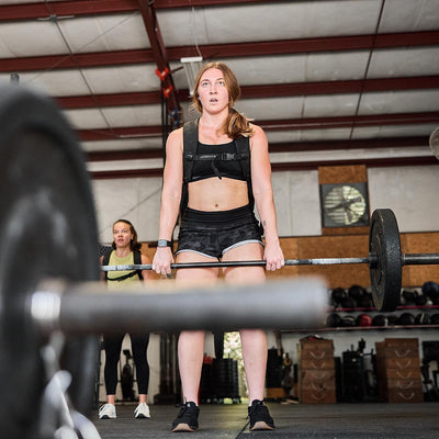 In a gym with high ceilings and organized equipment along the walls, a woman is focused on lifting a barbell. She’s wearing GORUCK's Women’s Training Shorts made with ToughStretch fabric, which ensure ease of movement. In the background, another person watches her workout.