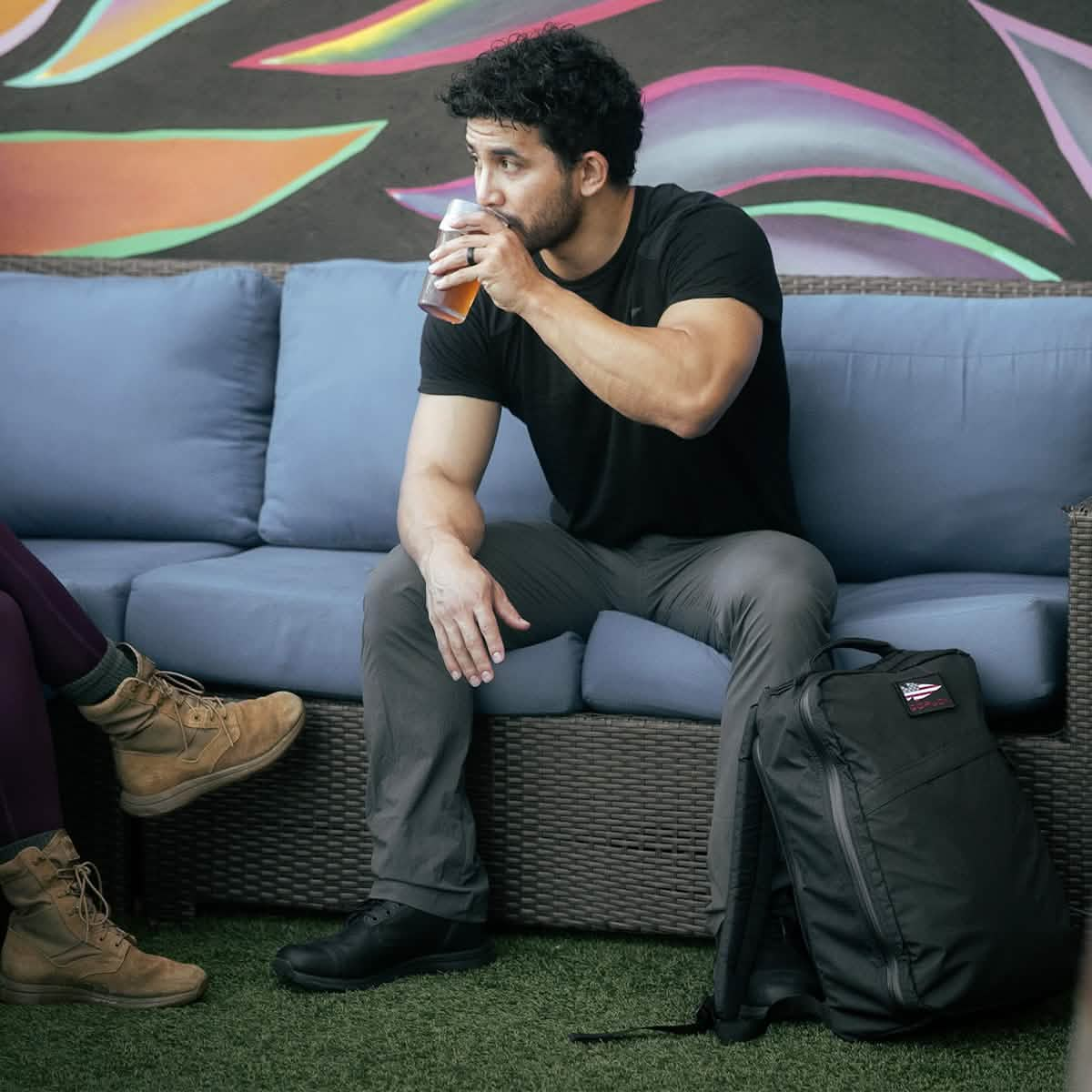 A person with curly hair relaxes on a blue sofa in an outdoor setting, sipping a drink. They're clad in a black shirt and gray pants, with the GORUCK GR1 USA - X-PAC rucksack resting at their feet. To the left, there is another person wearing boots, accompanied by a vibrant mural displayed in the background.