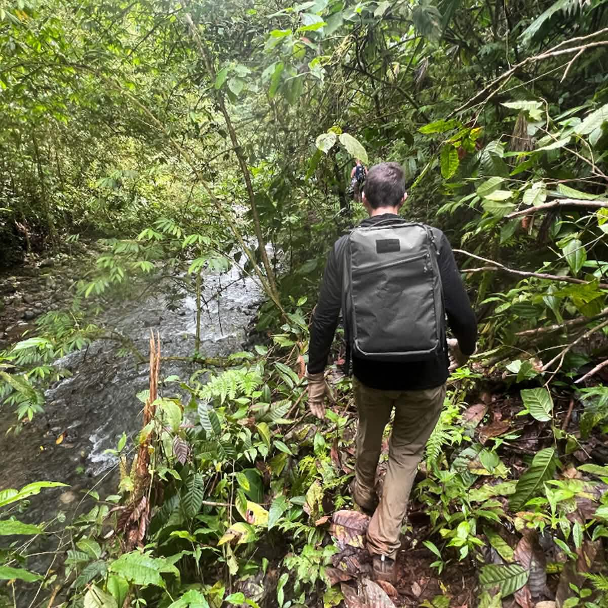 A person with a GORUCK GR1 USA - X-PAC backpack navigates through a dense, green forest next to a flowing stream. They are dressed in hiking-appropriate outdoor clothing and are enveloped by lush vegetation.