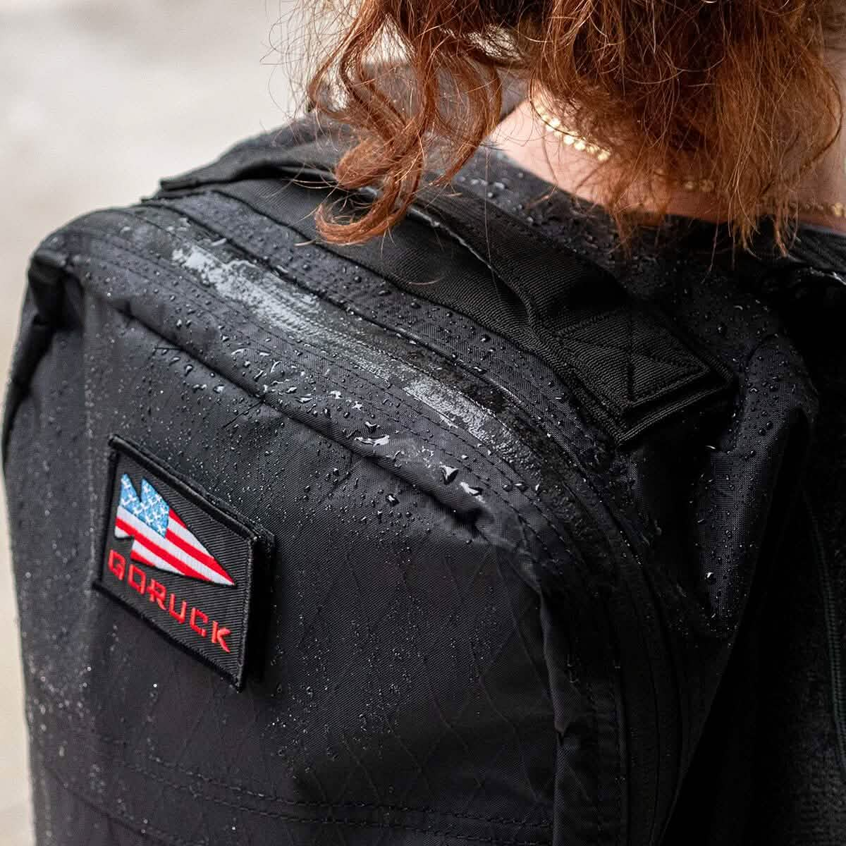 A person wearing a black GORUCK GR1 USA - X-PAC rucksack adorned with an American flag patch navigates the rain-soaked streets. The waterproof backpack glistens with raindrops, perfectly complementing the person’s curly hair.