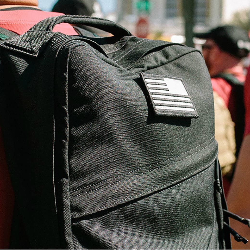 Close-up of a black backpack displaying the "Patch - Reflective Reverse Flag" by vendor-unknown, with an embroidered flag design and VELCRO backing, worn by someone in a crowd. The background is intentionally blurred to highlight various individuals and urban scenery in the distance.