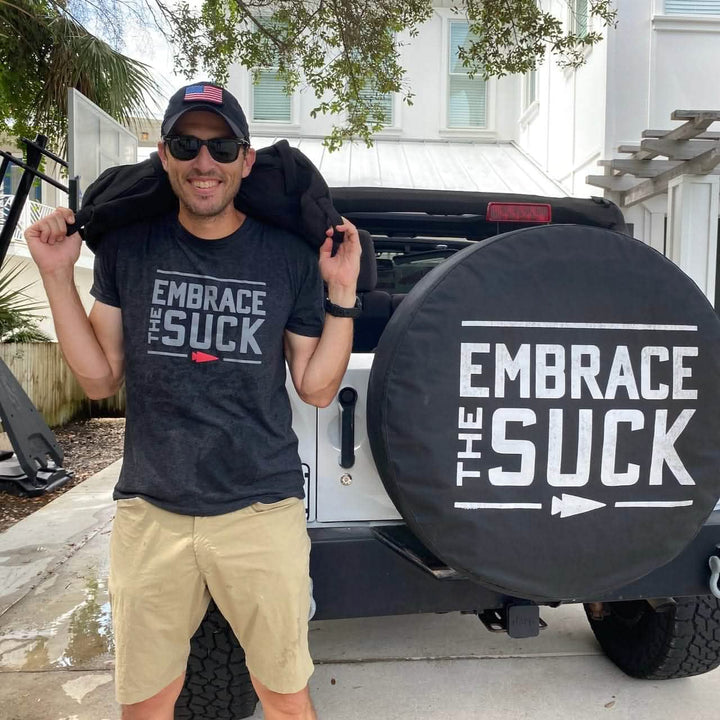 A person in a black cap and sunglasses stands next to a white Jeep, featuring a rear tire cover emblazoned with "Embrace the Suck"—an unofficial nod to USMC resilience. They're wearing an Embrace the Suck Tee - Tri-Blend by vendor-unknown and have a jacket draped over their shoulders.