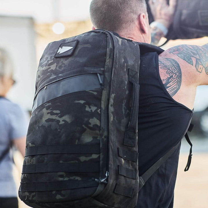 A person carrying a large, black camouflage backpack embellished with GORUCK's Patch - Reflective Spearhead ventures outdoors, with tattoos peeking from beneath their sleeveless shirt. In the background, another individual is visible, also carrying a backpack that features VELCRO closures for easy access.