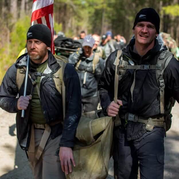 Two men, equipped for a cold weather rucking challenge, carry a bag along a dirt path through the wooded area. One holds a flag as they move with focus and determination, others closely following in this sunny GORUCK event with dappled light filtering through the trees. They are sporting the GORUCK Performance Beanie, which stands out in their gear.