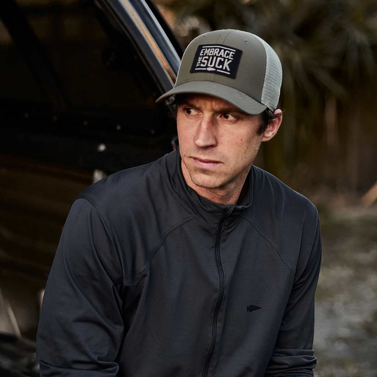 A man wearing a GORUCK Performance Trucker Hat - Embrace the Suck, featuring an adjustable velcro strap, sits looking to his left. The background is blurry, and he appears focused and contemplative.