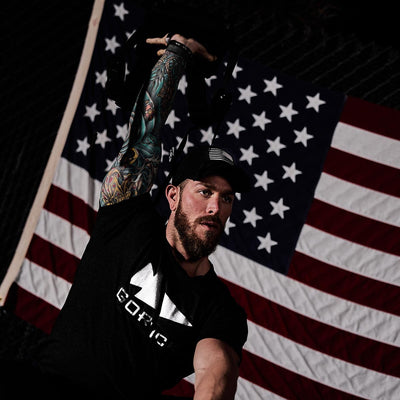 A man with tattoos on his left arm, wearing a GORUCK Reflective Tee - Tri-Blend and a matching cap, exercises with a kettlebell. An American flag hangs in the background, subtly reminiscent of special forces training.