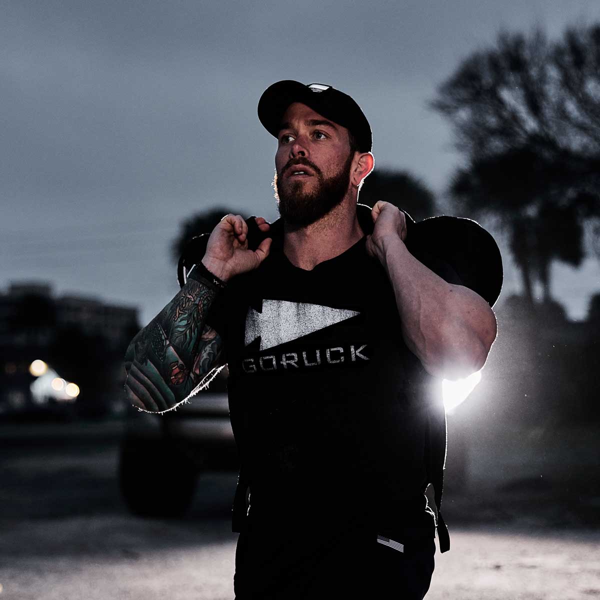 A man wearing a GORUCK Reflective Tee - Tri-Blend and a black cap carries a heavy backpack on his shoulders. With a tattoo on his left arm, he stands against a blurry urban background, vehicle headlights illuminating the scene behind him.