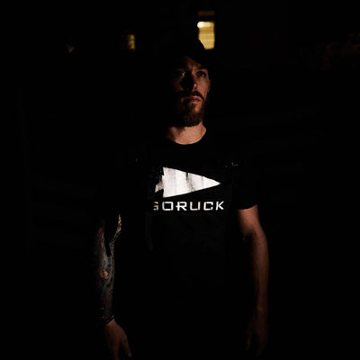 In a dimly lit setting, a bearded individual wears a black GORUCK Reflective Tee - Tri-Blend, embodying an aura of special forces coolness. A tattooed arm adds to the atmosphere, set against the subtle backdrop of what seems to be an American flag.