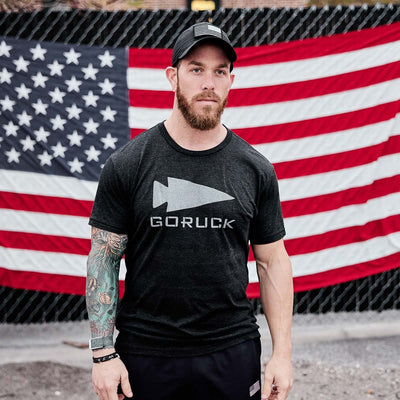 A man with a beard stands in front of a large American flag, wearing a black cap and the GORUCK Reflective Tee - Tri-Blend by vendor-unknown, featuring an arrowhead design reminiscent of special forces insignia. He has a sleeve tattoo on his right arm and looks directly at the camera.