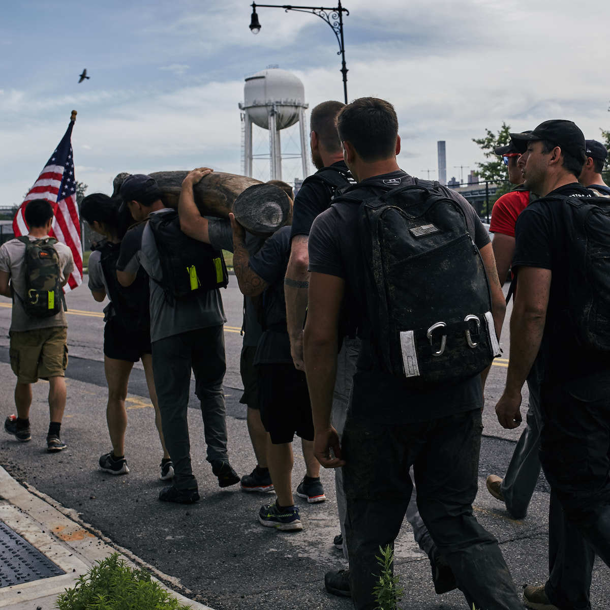 A group of individuals in reflective athletic gear, each featuring the Patch - Reflective Reverse Flag by vendor-unknown, carry a heavy log while walking along a street. One participant proudly holds up an American flag. In the background, there is a water tower and a streetlamp set against a cloudy sky.