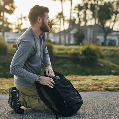 A man with a beard kneels on the pavement, dressed in a gray long-sleeve shirt, khaki pants, and black boots. He's adjusting a Patch - Reflective Reverse Flag from vendor-unknown on his black backpack. In the background are blurred greenery and houses beneath a cloudy sky.