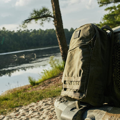 A green backpack featuring the vendor-unknown "Patch - Reflective Reverse Flag" patch rests on the edge of a vehicle near a tranquil lakeside, surrounded by trees and greenery under a partly cloudy sky.