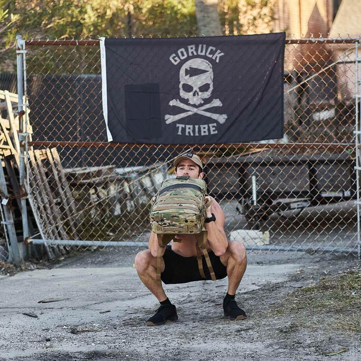 A person squats with a backpack outside, in front of a "Tribe" flag on a chain-link fence.