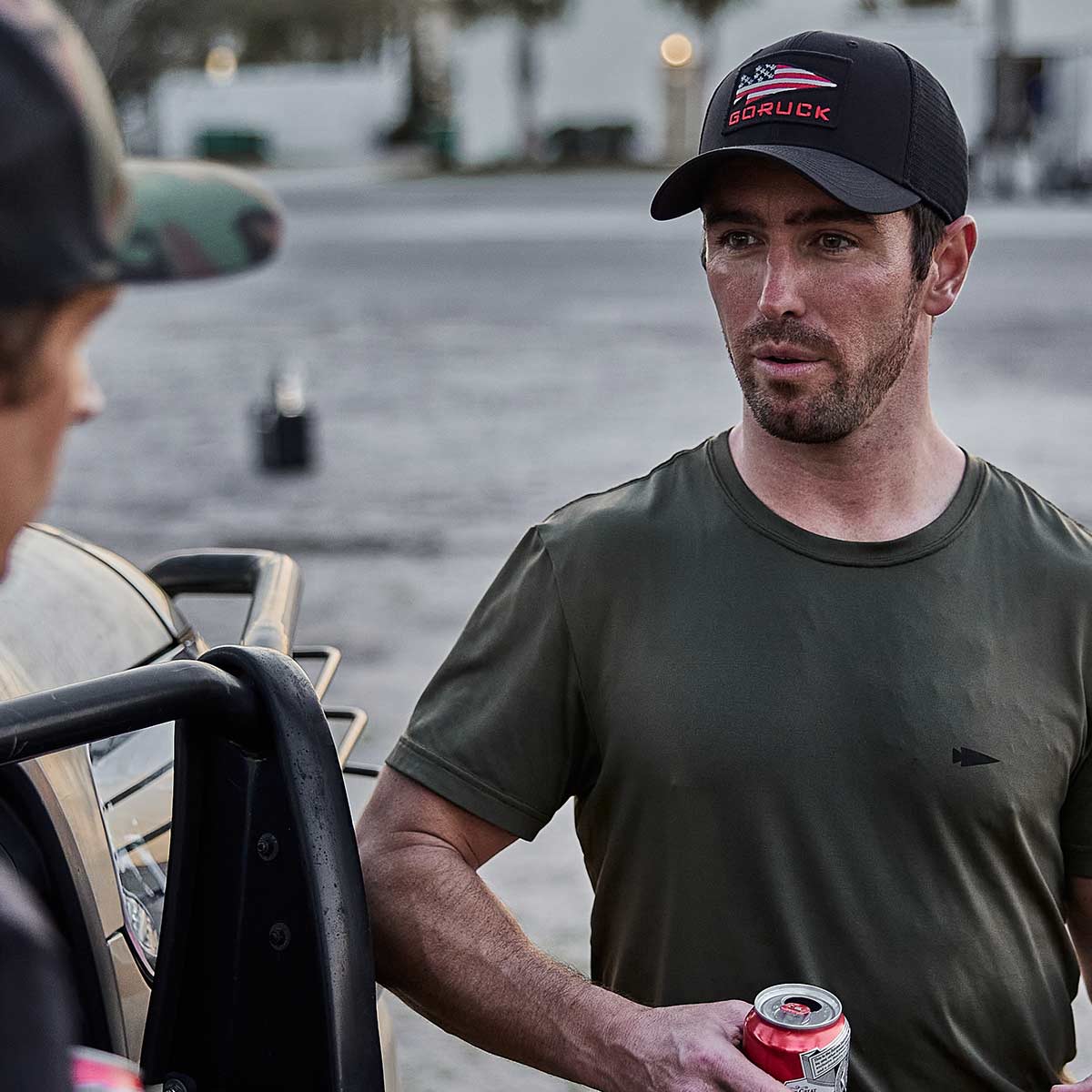 A man wearing a Performance Trucker Hat by GORUCK USA, which features an adjustable VELCRO® strap, and a green shirt holds a soda can while standing outdoors. He leans against a vehicle and talks to another person who is partially visible. The scene includes a blurred outdoor background.